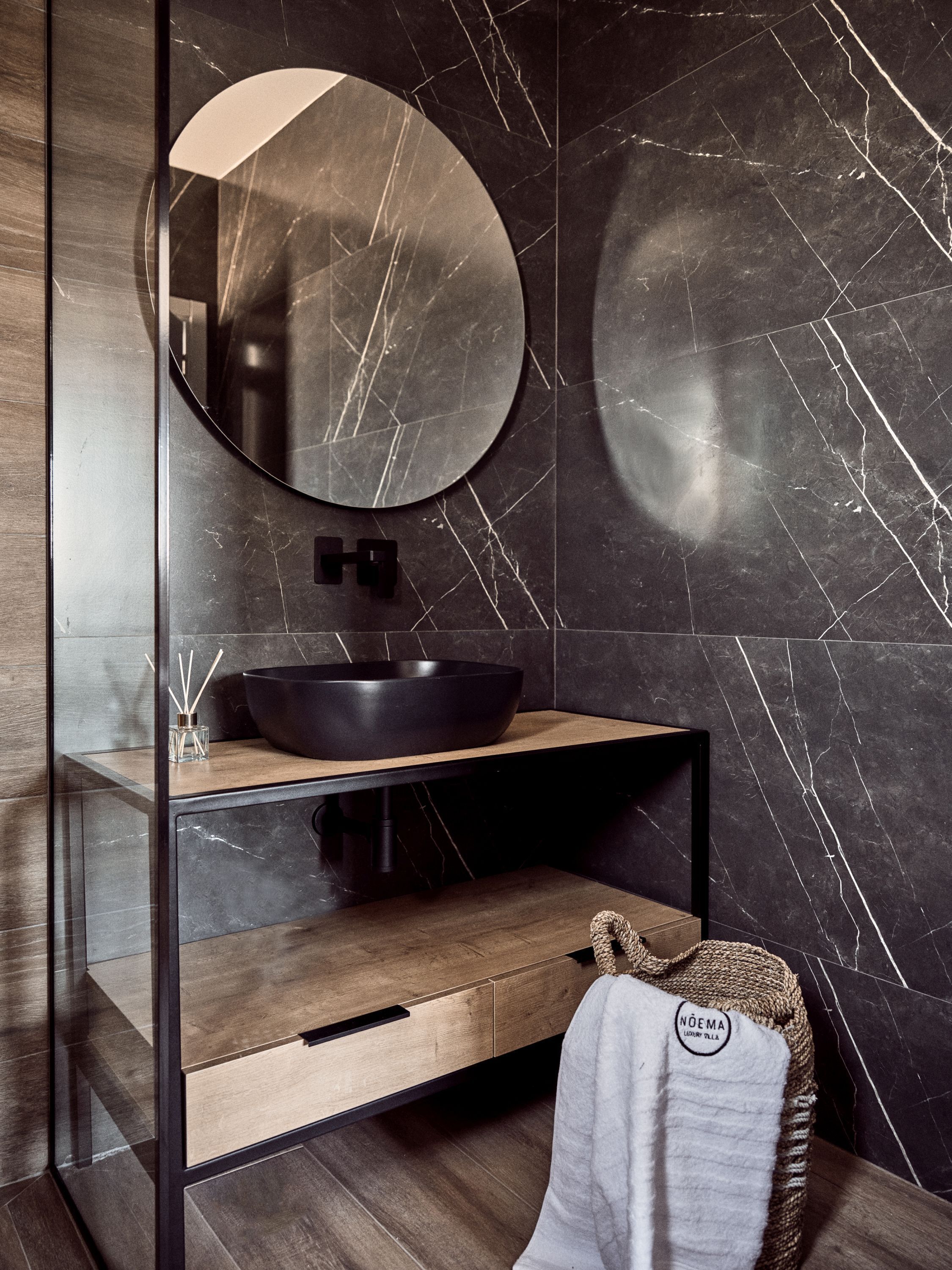 Une salle de bain moderne avec un mur en marbre noir, un miroir rond, un lavabo noir et un meuble-lavabo en bois.