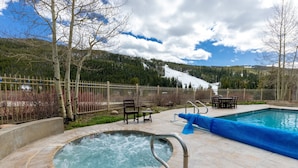 A hot tub with a metal railing, surrounded by a wooden fence, with a pool and outdoor furniture in the background.