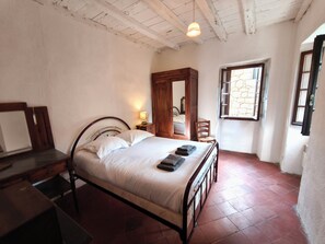 A bedroom with a bed, a chair, a wooden wardrobe, a nightstand, and a window with a stone wall.