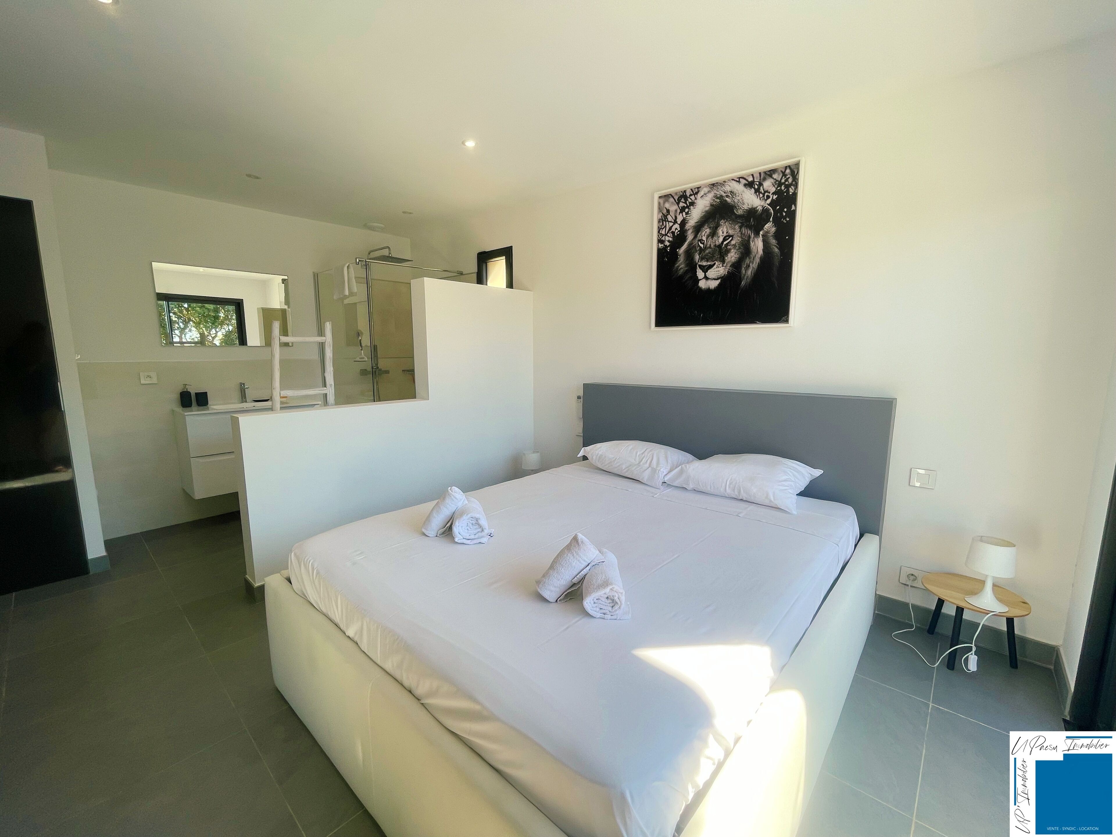 A modern bedroom with a large bed, a bathroom with a glass shower, and a painting on the wall.