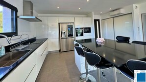 A modern kitchen with white cabinetry, a stainless steel refrigerator, a water dispenser, and a large island with bar stools. - Villa Ciste - 12/15pers - Heated swimming pool - Panoramic mountain view - (Zonza)