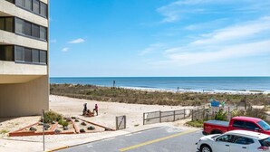 Una propiedad frente a la playa con vista al océano, un estacionamiento con una camioneta roja, y una persona caminando por la playa.