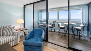 A room with a sofa, a blue armchair, a table with chairs, and a view of the ocean through large glass doors. - Gorgeous 2 BR/ 2 BA Beachfront Condo (Ocean City)