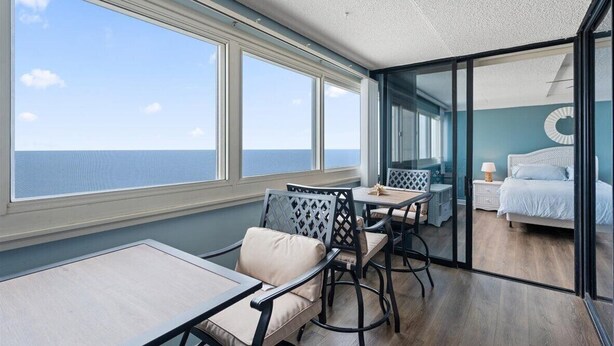 A room with a large window overlooking the sea, a bed with a circular wall decoration, and a dining area with chairs and a table. - Gorgeous 2 BR/ 2 BA Beachfront Condo (Ocean City)
