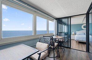 Una habitación con un amplio ventanal con vistas al mar, una cama con un adorno circular en la pared, y un comedor con sillas y una mesa.
