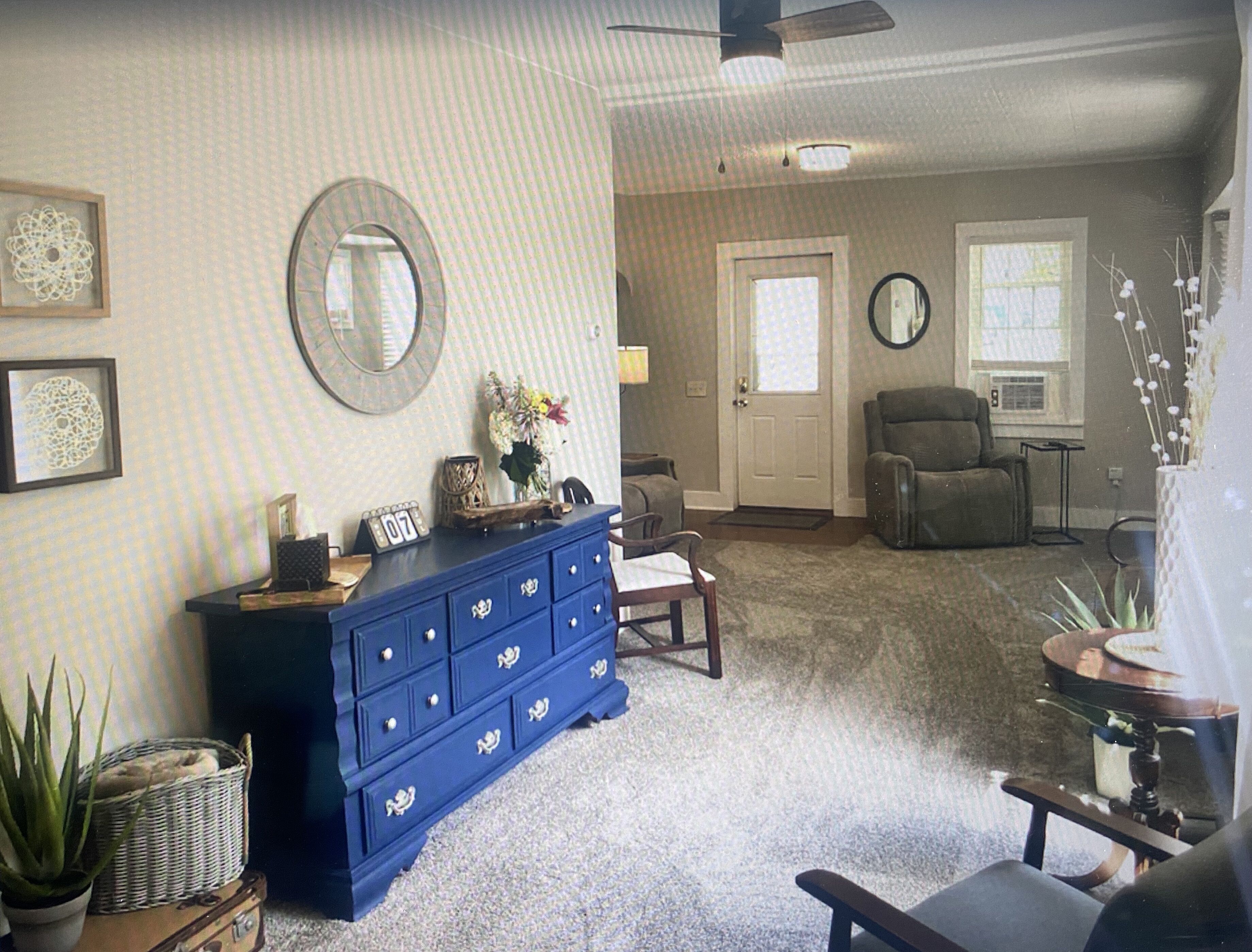 A living room with a blue dresser, a round mirror, a gray armchair, and a door with a window.