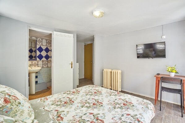 Quarto com edredom floral, televisão na parede, cadeira de madeira e radiador.