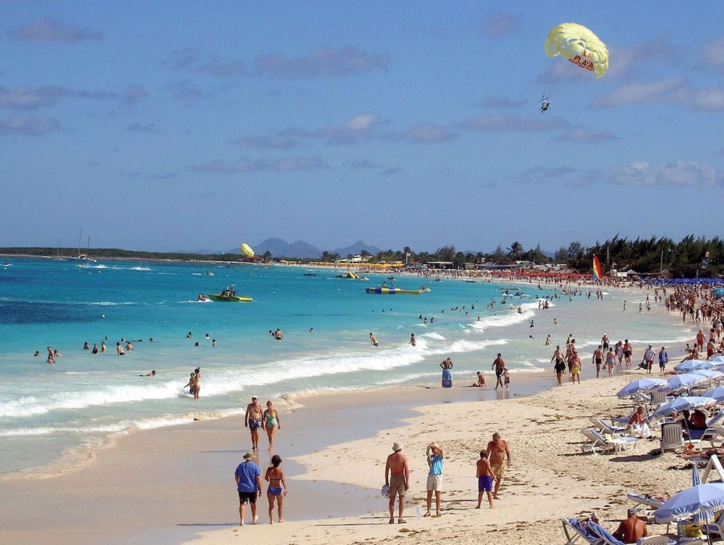 Une plage bondée où l’on voit des gens se promener, nager et faire du parachute ascensionnel.