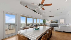 Un comedor amplio con una mesa grande, múltiples sillas, ventiladores de techo y vistas al océano a través de amplios ventanales.