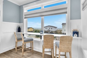 A room with a large window, wicker chairs, a white bench, and a view of houses and the ocean.