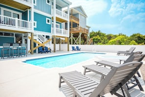 A swimming pool with lounge chairs, a wooden deck, and a multi-story building with balconies.