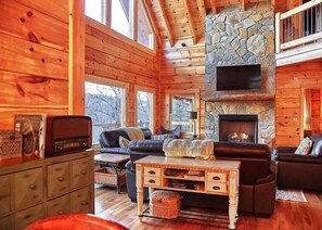 A cozy cabin interior with wooden walls, a stone fireplace, a flat-screen TV, and a leather sofa. - Gated Community, Hot Tub, Game Room (Lake Lure)