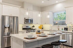 Una cocina moderna con electrodomésticos de acero inoxidable, una isla central con taburetes, iluminación colgante y un ventanal con persianas.