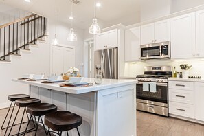 Cocina moderna con armarios blancos, electrodomésticos de acero inoxidable, una isla central con taburetes altos e iluminación colgante.
