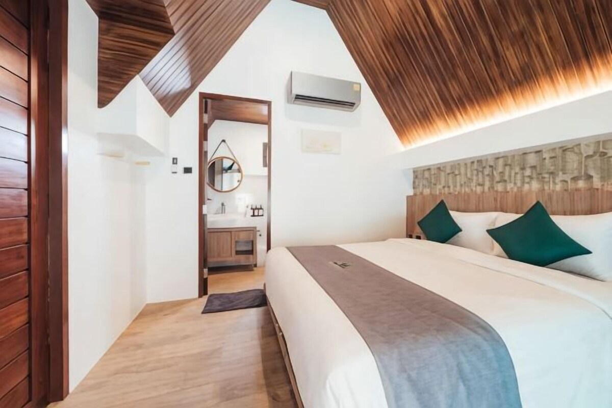 A bedroom with a wooden ceiling, a bed with green pillows, a bedside table, and a mirror.