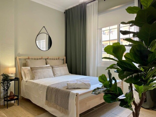 A neatly made bed with a wooden headboard, a bedside table with a lamp, and a large green plant in the corner.