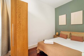 A bedroom with a wooden wardrobe, a bed with a beige blanket, two pillows, and a nightstand with a lamp.