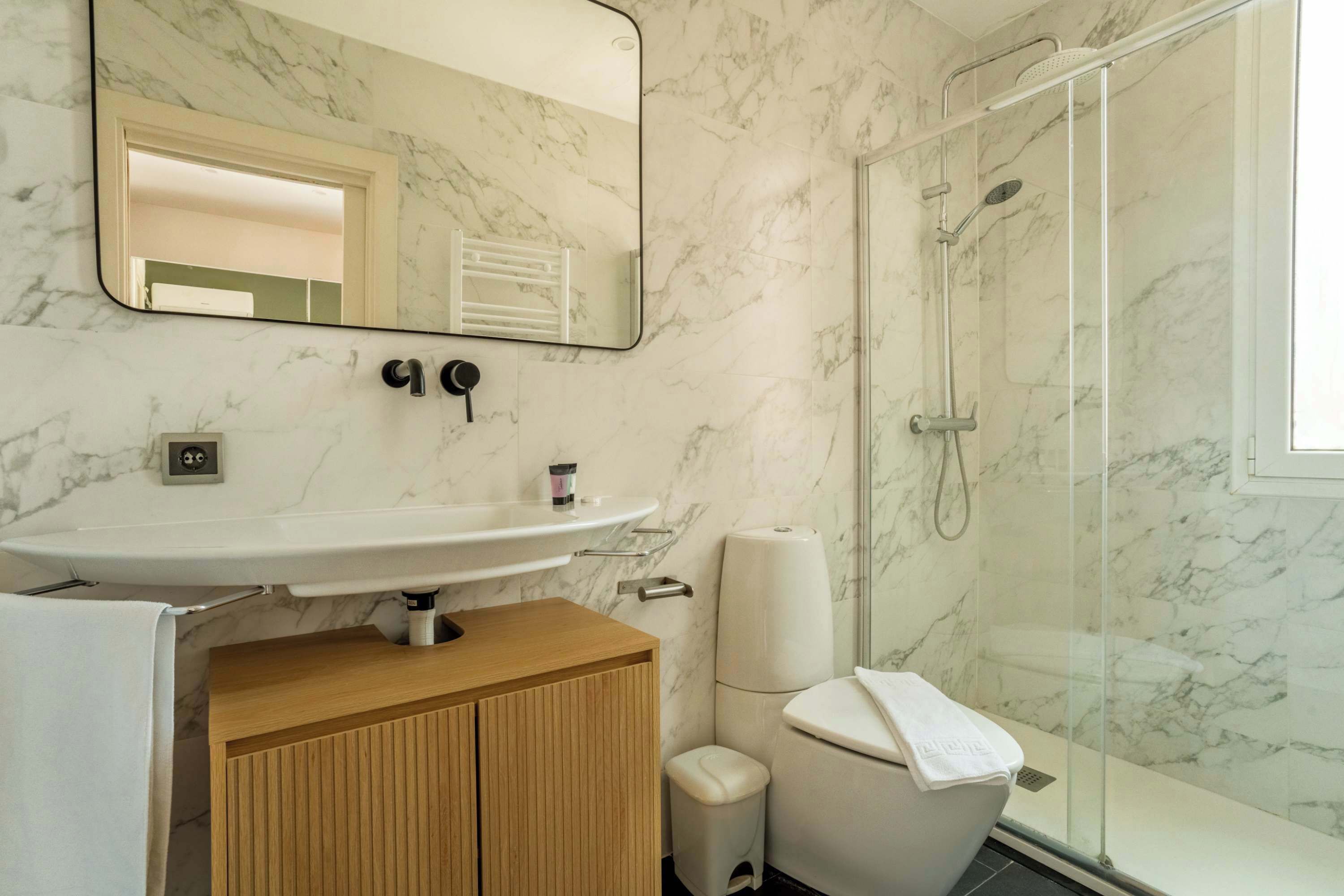 A bathroom with a marble wall, a white sink, a toilet, and a glass-enclosed shower.