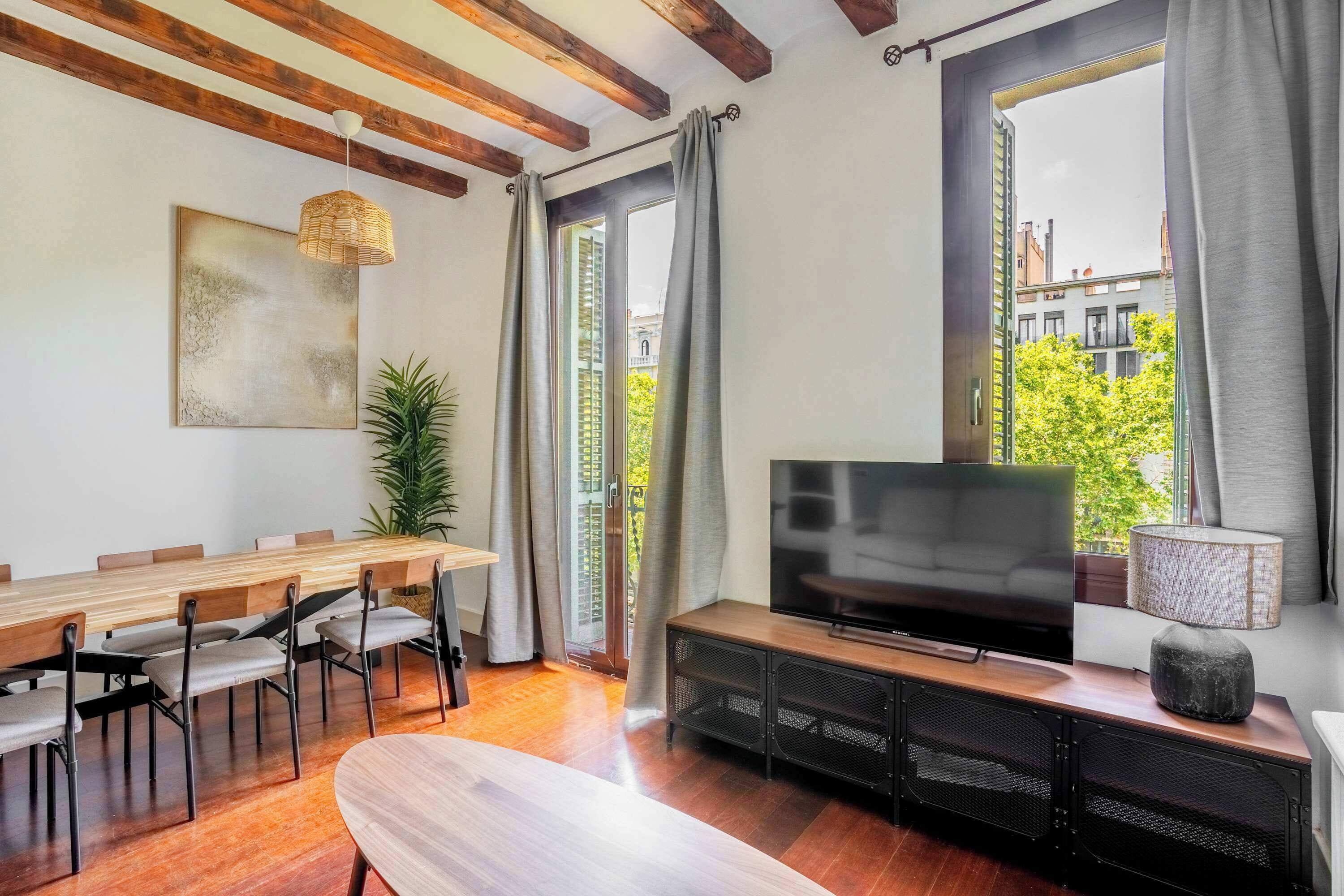 Ein modernes Wohnzimmer mit einem Holzesstisch, Stühlen, einem Flachbildfernseher und einem großen Fenster mit Vorhängen.