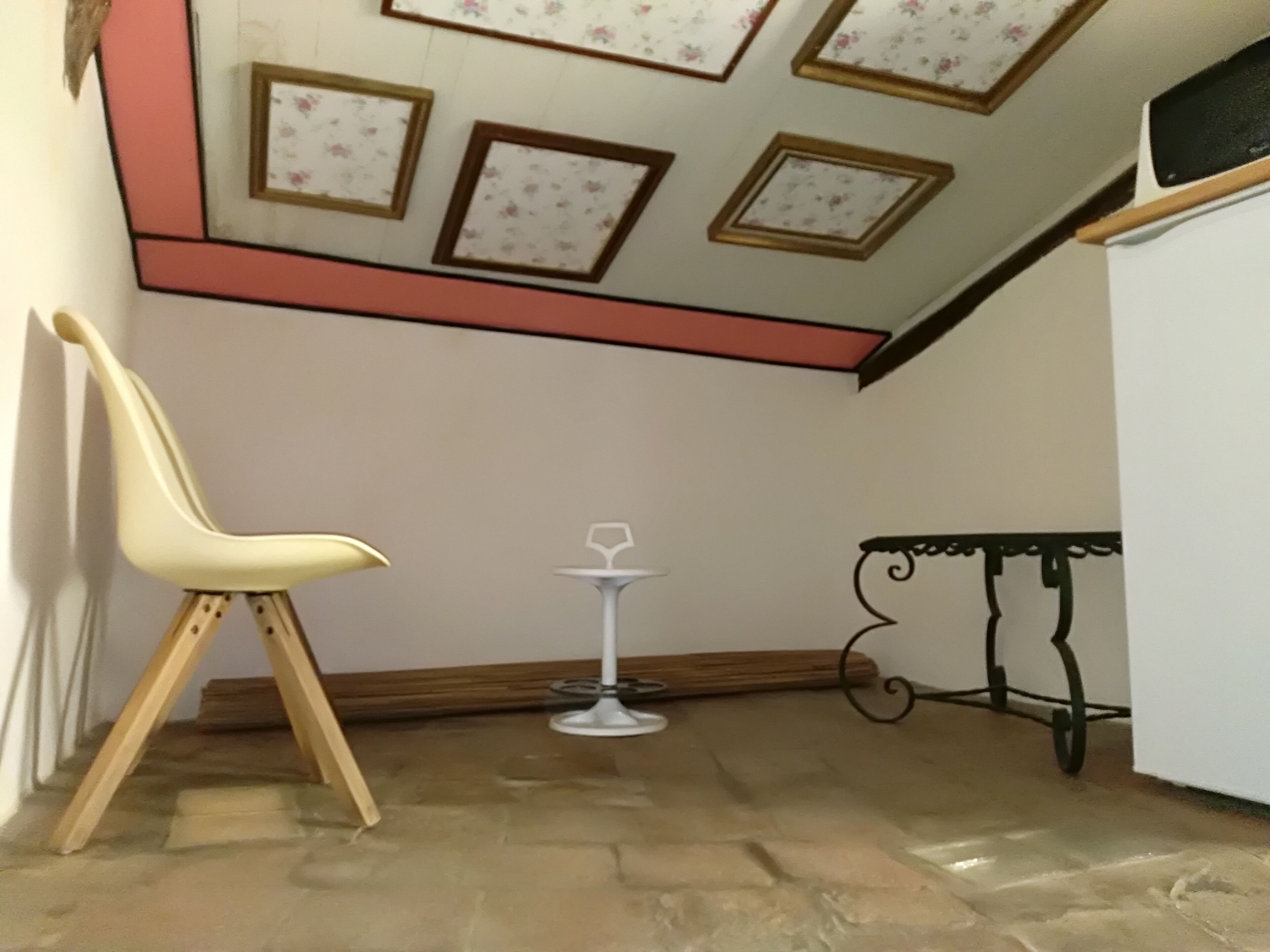 A corner of a room with a white chair, a small table with a wine glass, and a decorative ceiling with floral patterns.