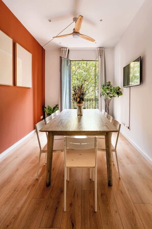A dining area with a wooden table, white chairs, a ceiling fan, and a view of greenery outside.