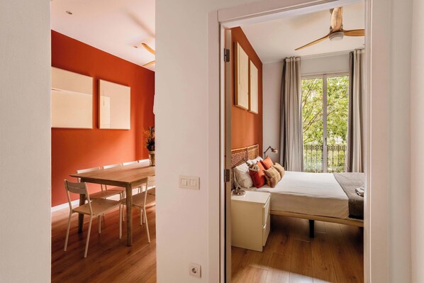 A room with a bed, bedside table, and a window with curtains. There is a wooden dining table with chairs in the adjacent room. - Beautiful 4bd 4ba apartment in Eixample District (Barcelona)