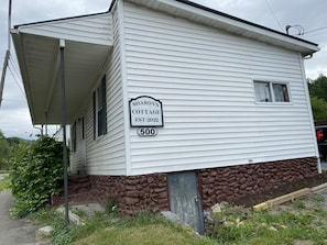 Una cabaña blanca con base de piedra, un porche y un cartel que indica que es Sharon's Cottage, establecida en 2022.