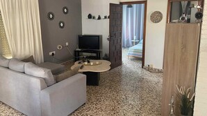 Ein Wohnzimmer mit grauem Sofa, einem Fernseher auf einem Standfuß, einem Couchtisch mit Getränken und einer Tür zu einem Schlafzimmer.