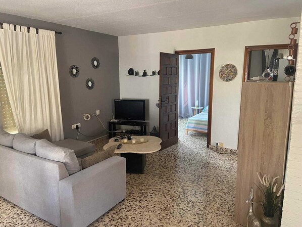A living room with a grey sofa, a TV on a stand, a coffee table with drinks, and a door leading to a bedroom.