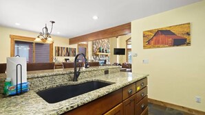 A kitchen with granite countertops, a sink, wooden cabinets, and a farmhouse-style painting on the wall.
