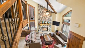 A spacious living room with a stone fireplace, wooden beams, and a large window.