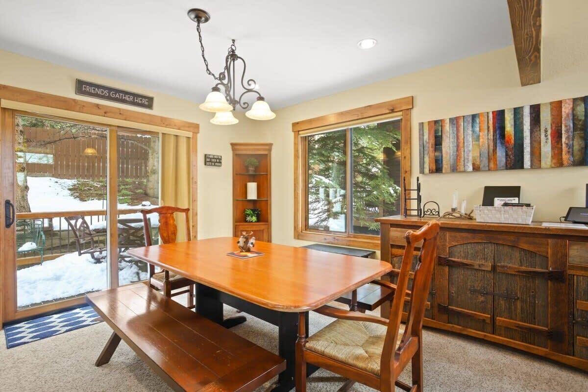 Un comedor con una mesa y sillas de madera, un banco, una araña y vistas amplias a través de grandes ventanales.