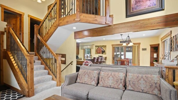 A spacious living room with a wooden staircase, a gray sofa, and a dining area in the background.