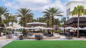 An outdoor seating area with tables and umbrellas, surrounded by palm trees and a building with a sign.
