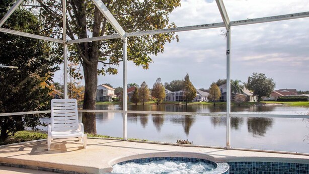 A pool area with a hot tub, a white lounge chair, and a view of a lake with houses in the background.