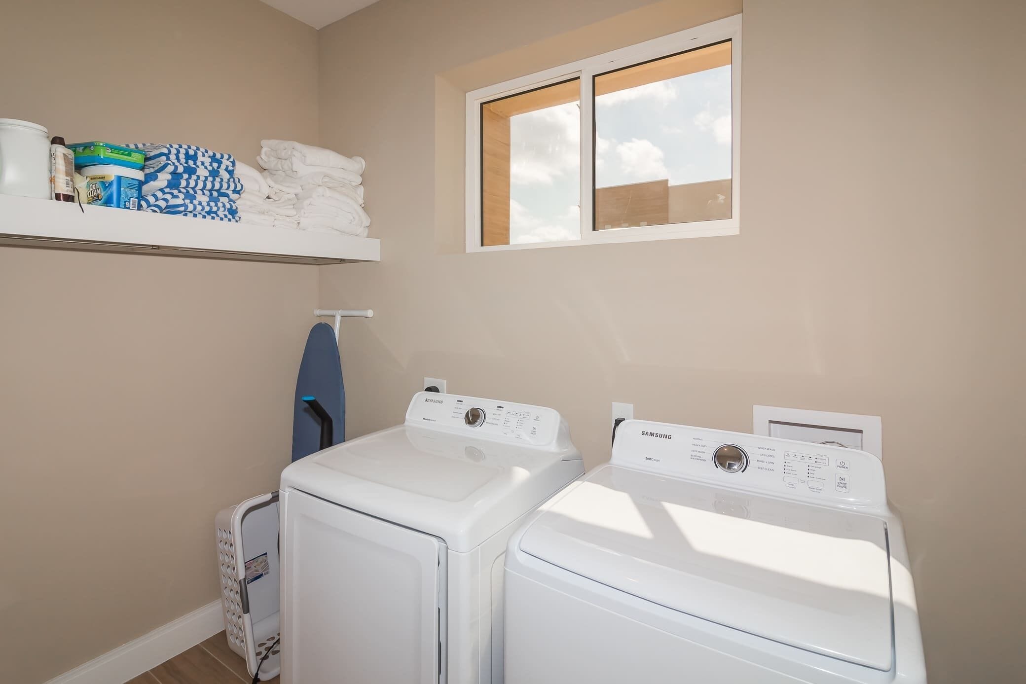 A laundry room with two white front-loading washing machines, a shelf with folded towels and laundry supplies, and a window with a view of the sky.