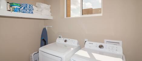 A laundry room with two white front-loading washing machines, a shelf with folded towels and laundry supplies, and a window with a view of the sky.