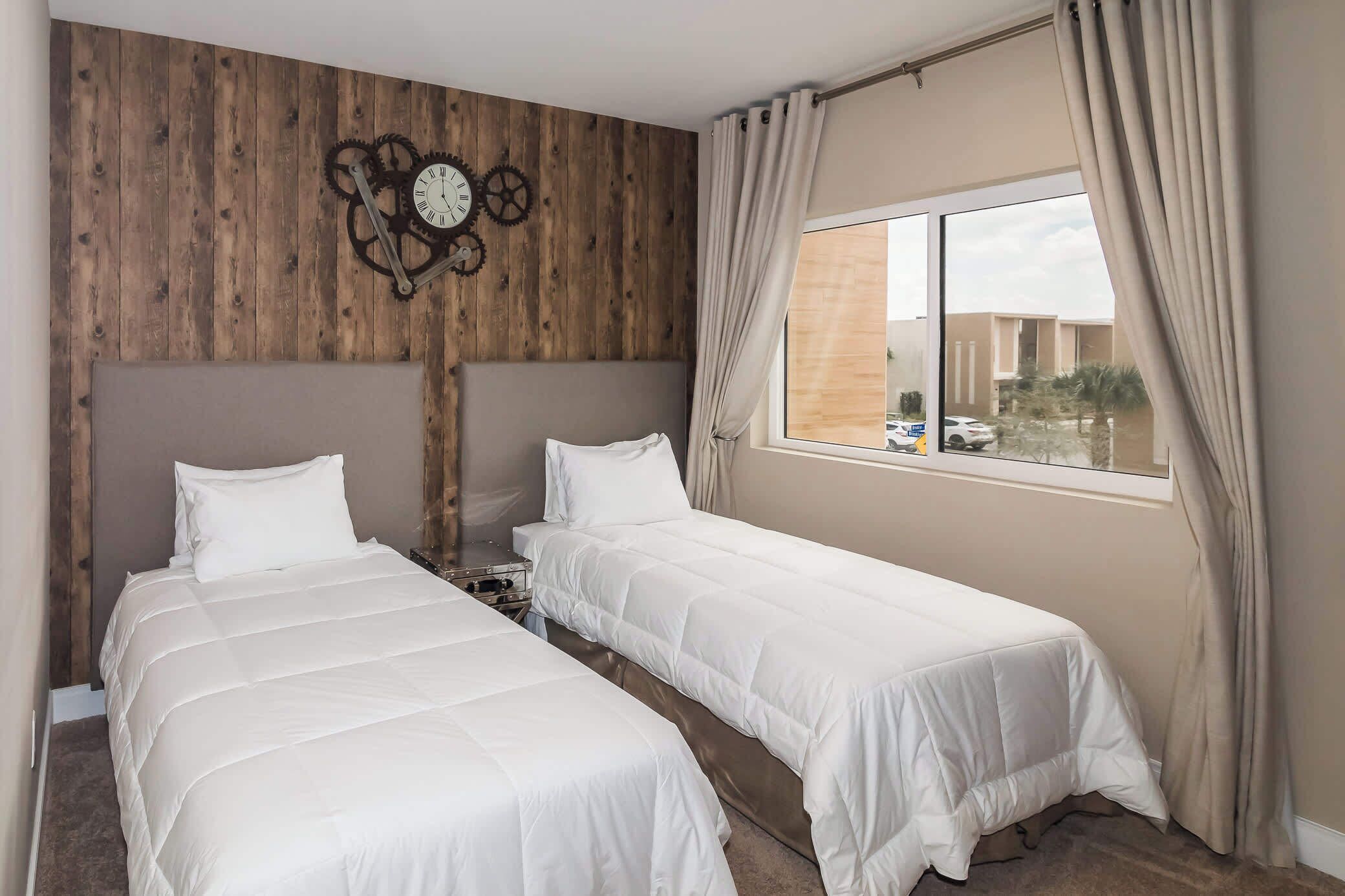 Une chambre avec deux lits, un mur en bois, une horloge et une fenêtre avec des rideaux.