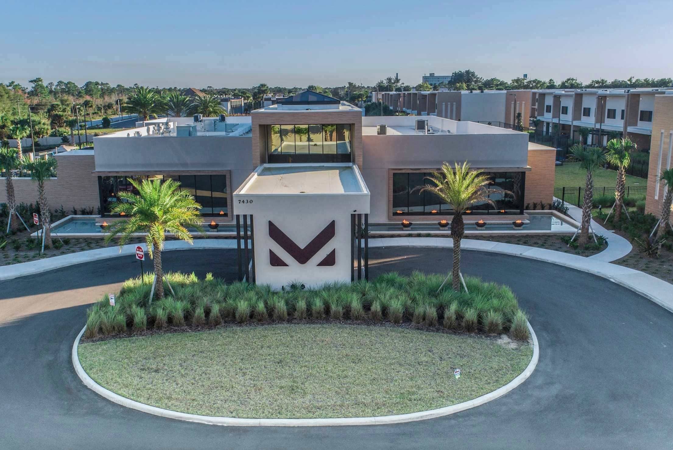 A modern building with a large entrance, a parking area, and a landscaped garden with palm trees.