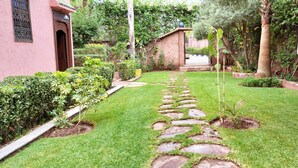 A garden with a stone pathway, lush greenery, and a pink building with a decorative door. - JNANE GHMATE (marrakech)