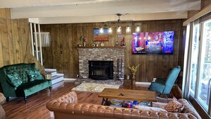 A cozy living room with a fireplace, a green sofa, a brown leather couch, and a television mounted on the wall. - 3 bedroom dream home 🏡 Big Bear Lake CA / Running Springs/Lake Arrowhead. (Running Springs)
