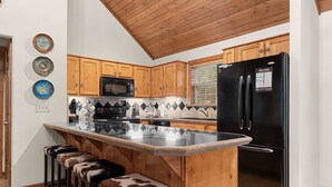 Cocina con armarios de madera, un refrigerador negro, un microondas y un taburete con asiento de cuero de vaca.