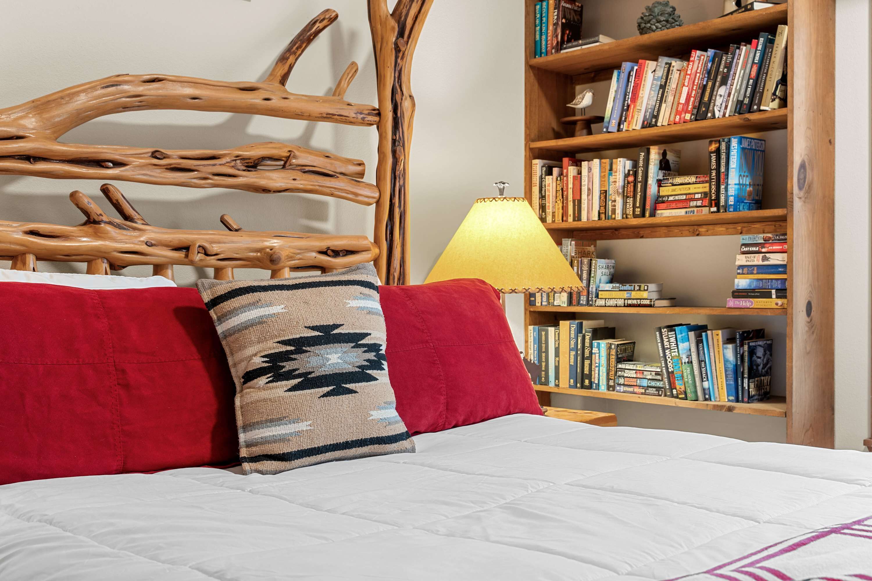 Een bed met een houten hoofdbord, een rood kussen, een kussen met een patroon, en een boekenplank vol boeken.