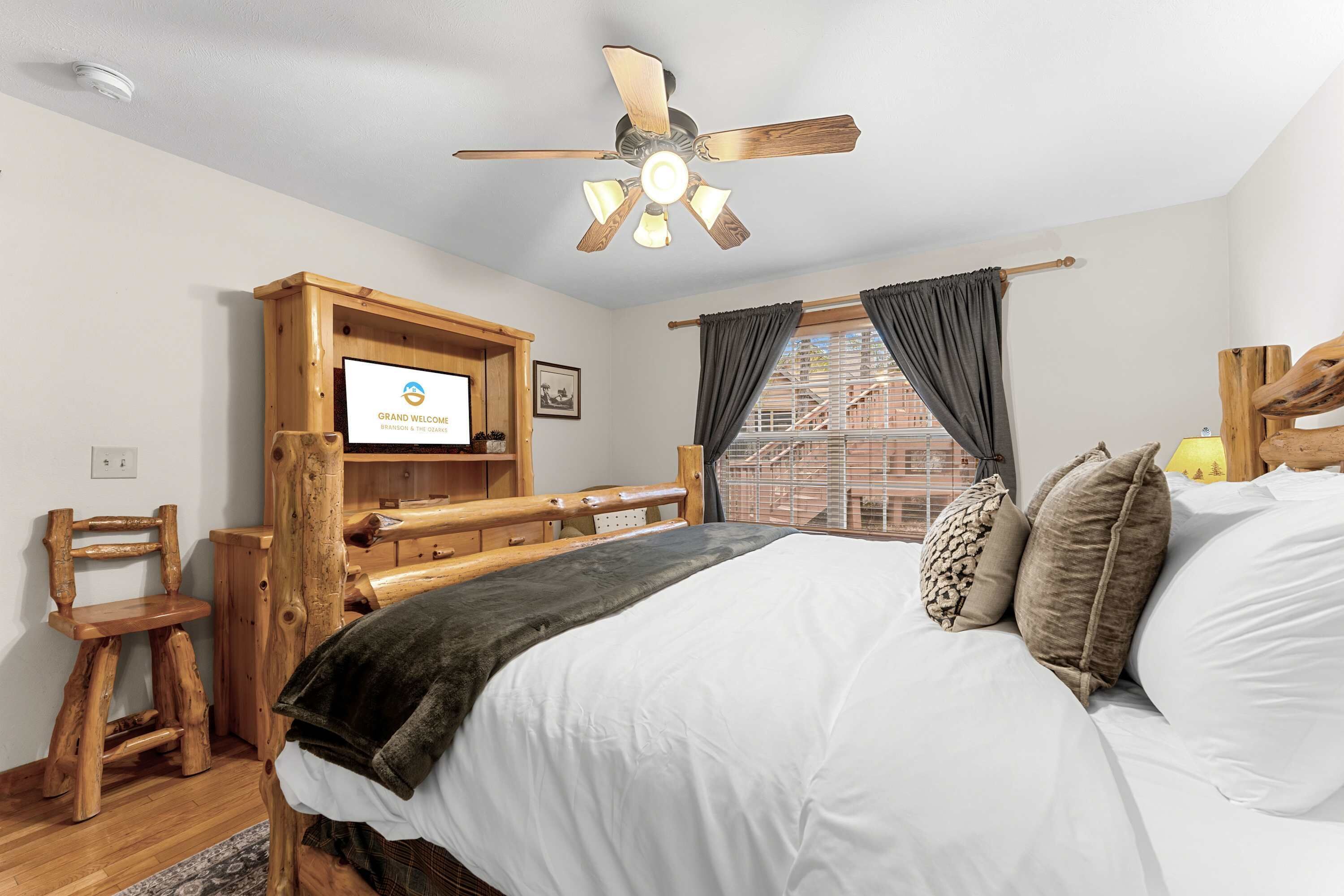 A bedroom with a bed, wooden headboard, a ceiling fan, a TV mounted on a wooden cabinet, and a window with curtains.