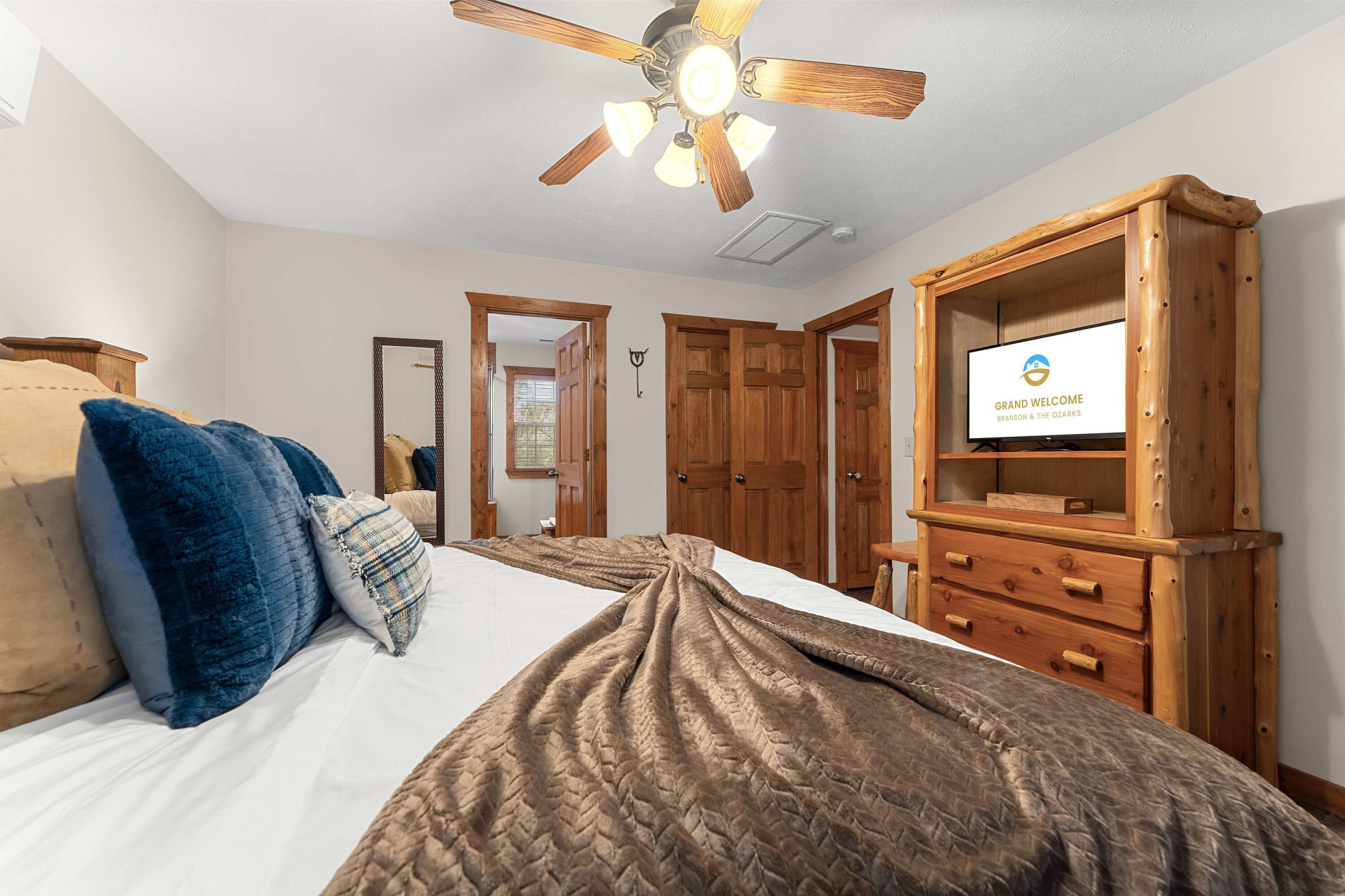 A bedroom with a bed, pillows, a wooden dresser, a ceiling fan, and a television displaying 'GRAND WELCOME.'