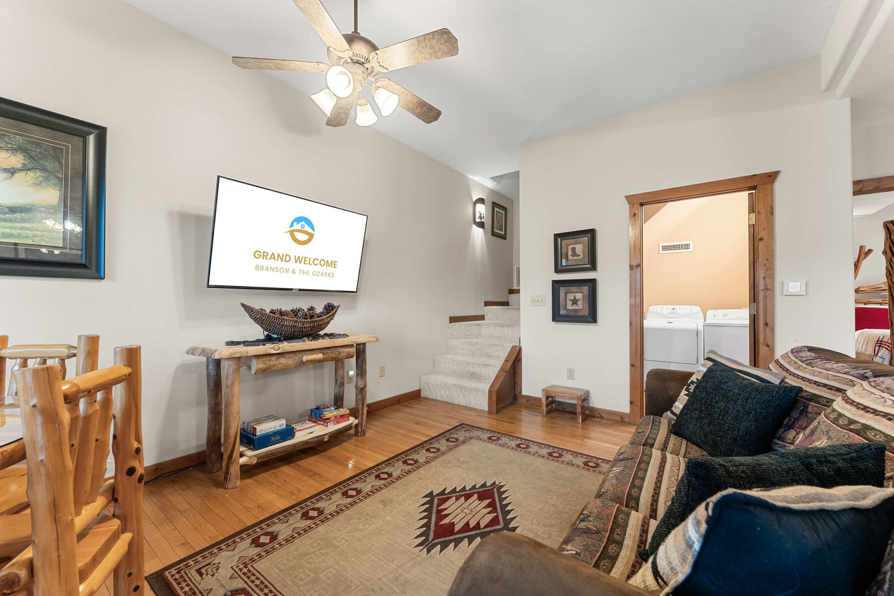 A living room with a television displaying 'Grand Welcome,' a ceiling fan, wooden furniture, a staircase, and a view into a laundry room.