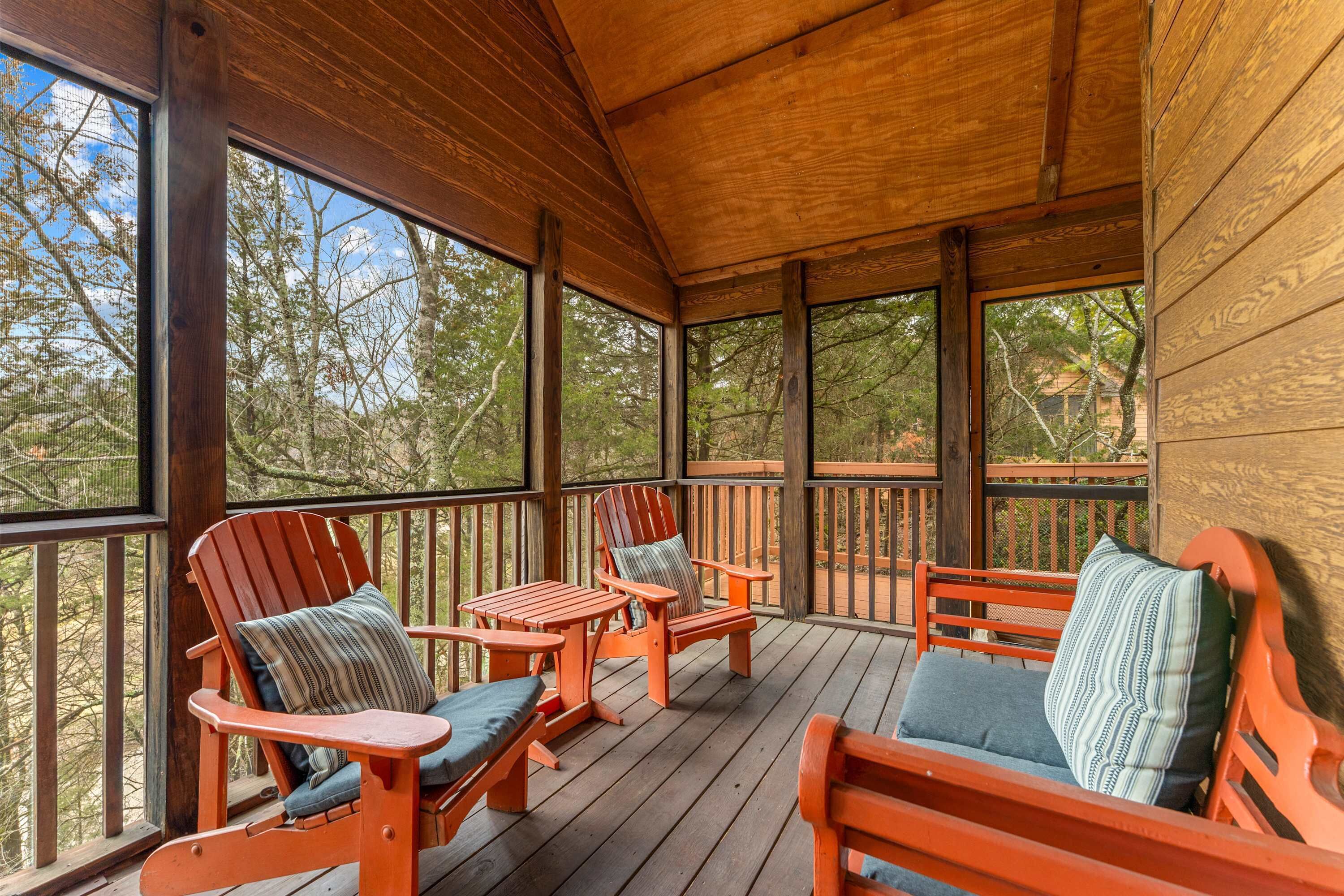 A wooden deck with Adirondack chairs and a small table, large windows, and a view of trees.