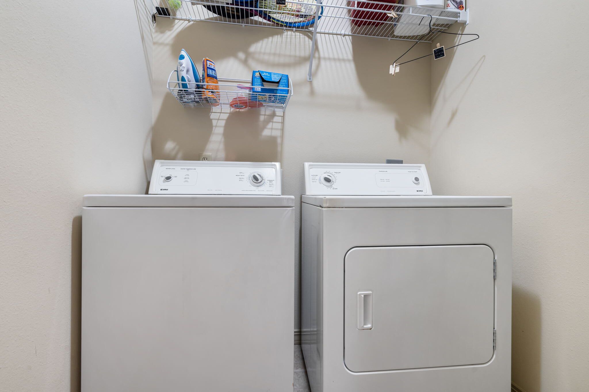Two front-loading washing machines in a laundry area with a shelf above holding various laundry supplies.