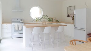 Une cuisine moderne avec des meubles blancs, un îlot central doté de tabourets, un four encastré et une grande fenêtre laissant entrer la lumière naturelle.