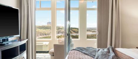 A hotel room with a bed, a TV, a desk, and a view of a cityscape through large windows.